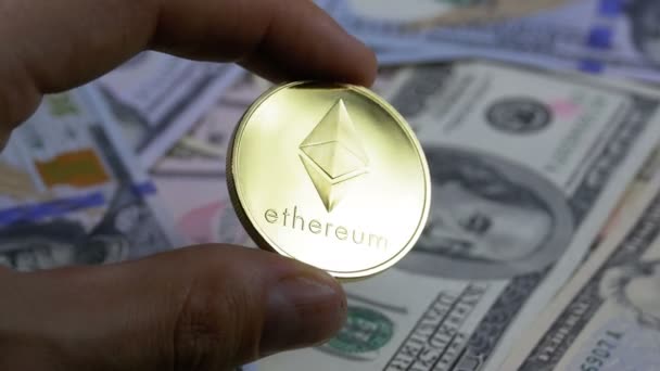 Χέρι με δάχτυλα κρατά ένα χρυσό νόμισμα Ethereum, Eth σε φόντο με τους λογαριασμούς δολαρίων — Αρχείο Βίντεο