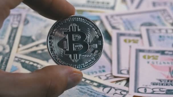 Tangan di Fingers Holds a Silver Coin Bitcoin, BTC on a Background with Bills of Dollars — Stok Video