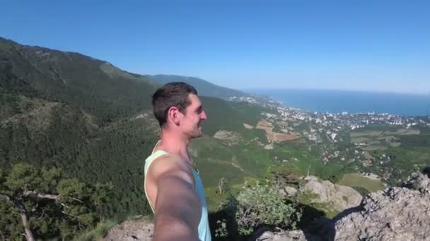 Man traveler at the top of a rocky mountain with Cross smiles, rejoices and rotates the camera around him — Stock Video