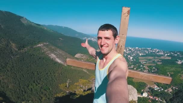 Fröhlicher junger Mann hebt die Hände hoch oben auf dem großen felsigen Berg mit Kreuz — Stockvideo