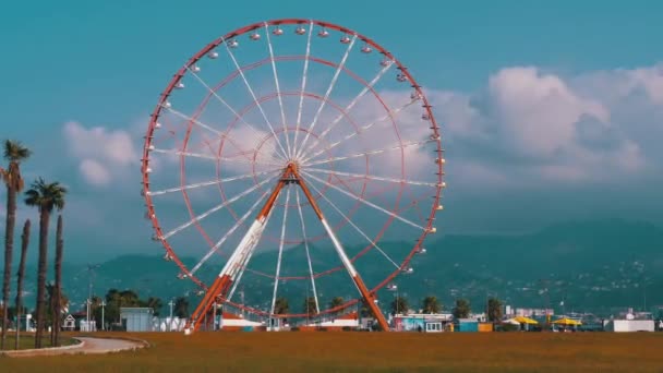 Колесо обозрения против голубого неба с облаками возле пальм в курортном городке, солнечный день — стоковое видео