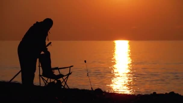 Silhueta de um pescador e cadeira de pesca ao pôr do sol caminho pelo mar . — Vídeo de Stock