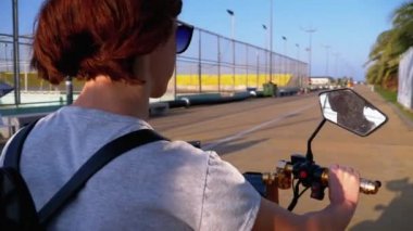Kadın, tatil köyünde Palmiye ağaçlarıyla Kırmızı Bisiklet Yolunda Elektrikli Scooter sürüyor.
