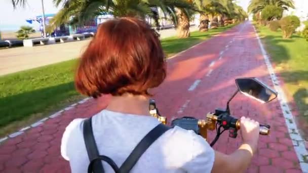 Γυναίκα Οδηγεί ένα ηλεκτρικό σκούτερ σε ένα κόκκινο μονοπάτι ποδήλατο με φοίνικες στην πόλη Resort — Αρχείο Βίντεο