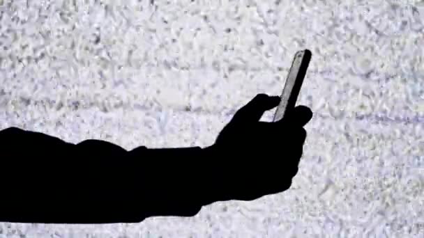 Силуэт руки, держащей смартфон на фоне экрана телевизора с белым статическим шумом — стоковое видео