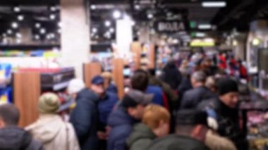 Bir süpermarketteki alıcılar sıra sıra mal arasında yürürler. Satıcılar rafların arkasında duruyor.