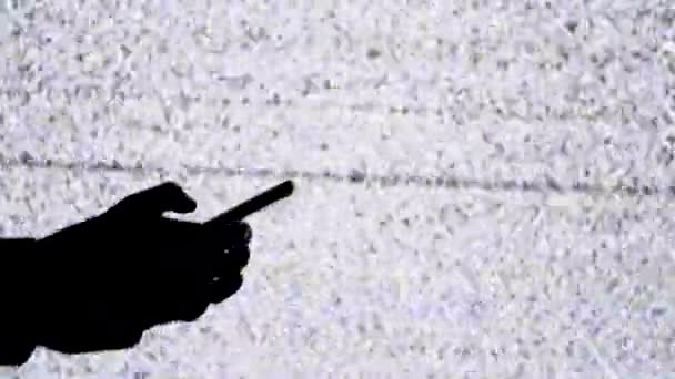 Силуэт руки, держащей смартфон на фоне экрана телевизора с белым статическим шумом — стоковое видео