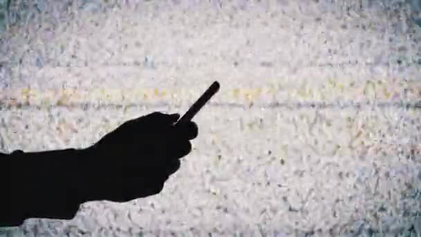 Σιλουέτα ενός χεριού κρατώντας ένα Smartphone στην οθόνη τηλεόρασης φόντο με λευκό στατικό θόρυβο — Αρχείο Βίντεο