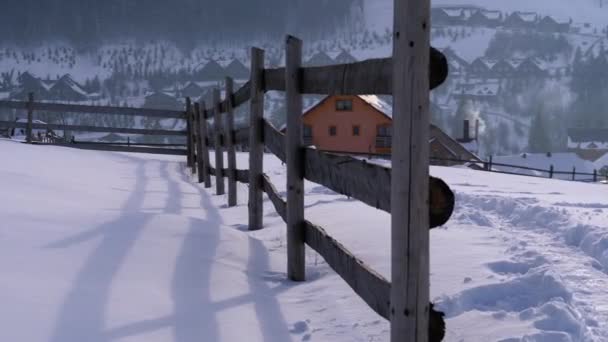 Cena rural de inverno com cerca de madeira abandonada velha e casa de madeira nevada e montanhas — Vídeo de Stock