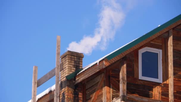 Humo de una chimenea de ladrillo en un tejado de casa de madera en invierno contra un cielo azul . — Vídeos de Stock