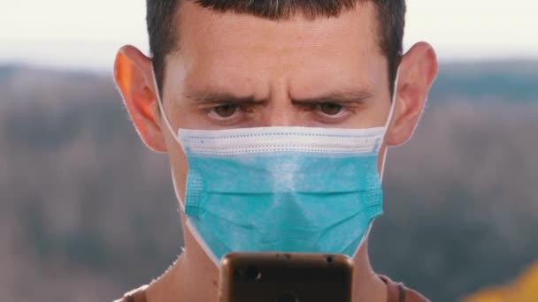 Человек в защитной маске смотрит на смартфон, держащий его в руках на улице — стоковое видео