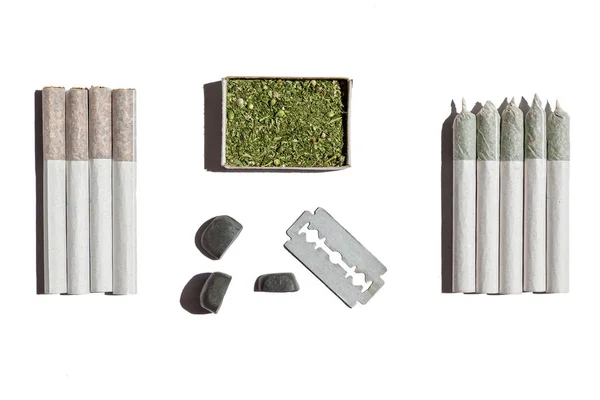 Quatre cigarettes roulées avec du tabac, cinq avec du chanvre, boîte avec du cannabis, lame et pierres posées sur la table blanche . — Photo