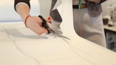 Kadınların elleri beyaz bir masanın üzerinde terzi makası ile kağıdın desenini kesiyor. Kumaş, eller, dikiş, tasarım