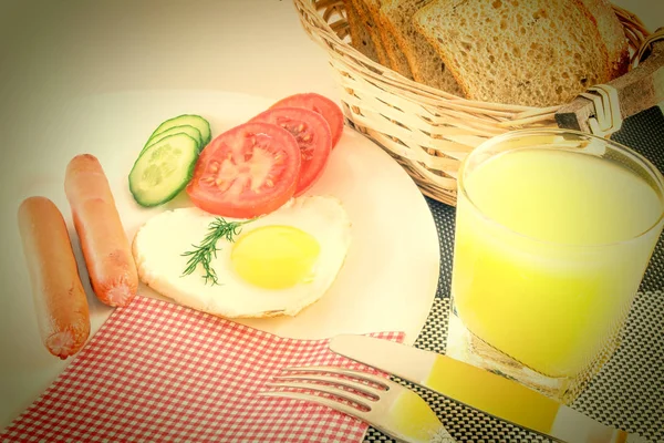 Breakfast on a table, fried egg in a heart-shaped fried sausages, fresh sliced vegetables cucumbers and tomatoes, juice, sliced bread, knife and fork, napkin. Old grunge style. — Stock Photo, Image