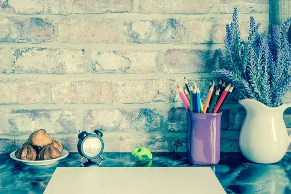 Colored pencils in a mug, vase of lavender flowers, clock, plate of cakes, white paper on a table against a brick vintage wall. Old retro vintage style photo.