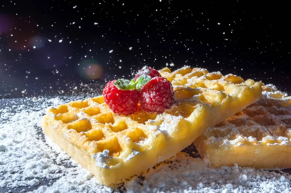 Waffles torradas doces com framboesas e um raminho de folhas de hortelã em cima e peneiração verter açúcar em pó na macro luz solar close-up em um fundo preto — Fotografia de Stock