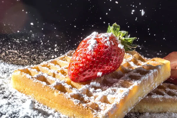 Doces waffles torradas com morango com folhas em cima e peneirando verter açúcar em pó na luz solar macro close-up em um fundo preto — Fotografia de Stock