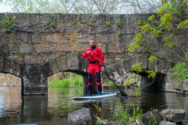 一名身穿红色干衣的男子沿着一座石桥附近的河边登上了一辆Sup牌 免版税图库照片