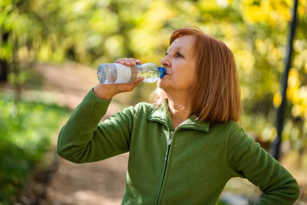 Senior woman is drinking water after exercising in park.