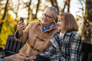 Yaşlı çift parkta bankta oturuyor ve sonbaharın tadını çıkarıyor..