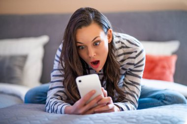 Evinde yatakta uzanan ve akıllı telefon kullanan bir kadın..