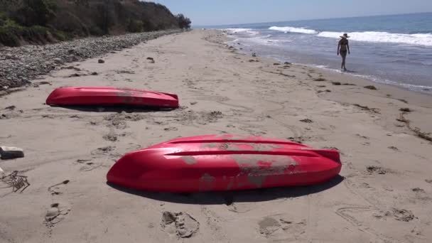 一个戴着帽子的女人走在南加州的海滩上 她的前景是一双红皮划艇 — 图库视频影像