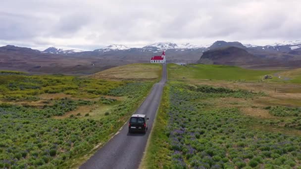 アイスランドの山の中の丘の上の教会まで車で行く黒いキャンパーバンの上に空中 — ストック動画