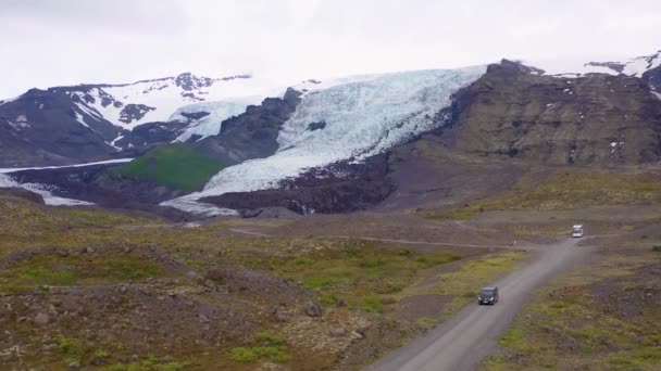 アイスランドの山の中にある遠くの氷河まで運転する黒いキャンパーバンの上の空中 — ストック動画