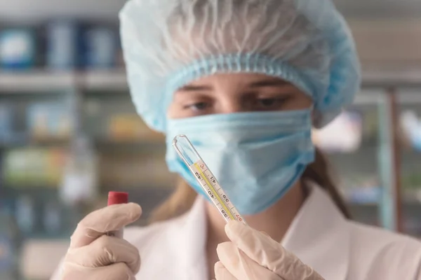 Young female pharmacist in protective medical mask and cap with thermometer. Health protection from the virus concept. Image with selective focus