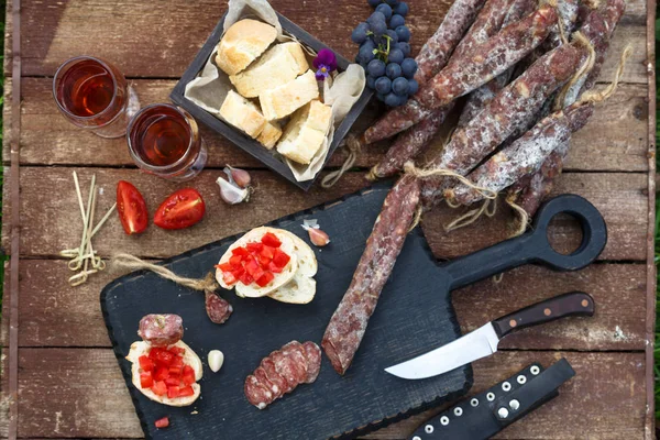 Wine and appetizers set. Bread, dry cured pork meat sausage, tomato on rustic wooden serving board and red wine in glass over rustic background, top view — Stock Photo, Image