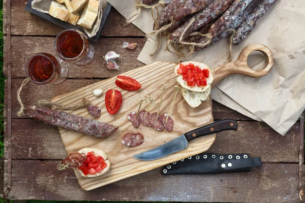 Brot, Wurst, Rotwein, Glas, Schneidebrett und Messer auf einem Holztisch für einen Imbiss in der Natur arrangiert. — Stockfoto