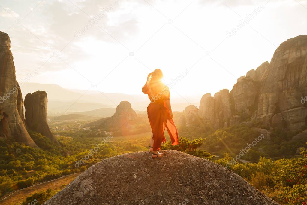 Young woman in red dress looking at majestic sunset in Meteora valley, Greece