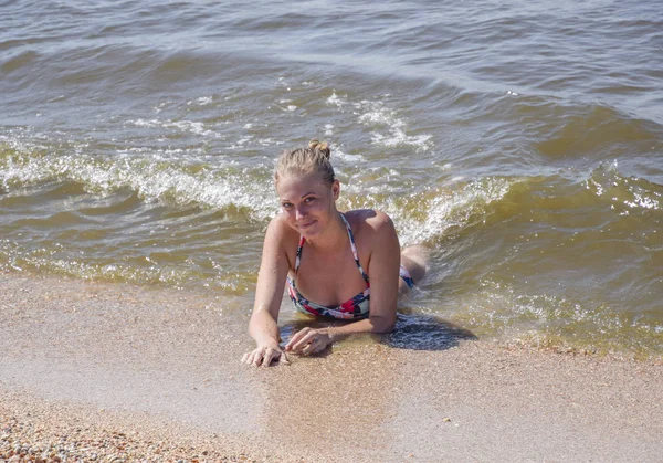 Blond meisje in een bikini, liggend op het strand en de golven splash daarop. Mooie jonge vrouw in een kleurrijke bikini op zee achtergrond — Stockfoto