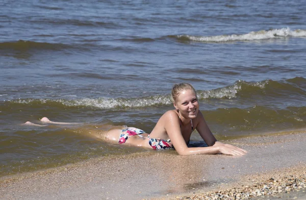 Ξανθό κορίτσι σε ένα μπικίνι που βρίσκεται πάνω στην παραλία και το splash κύματα σε αυτό. Όμορφη νεαρή γυναίκα σε ένα πολύχρωμο μπικίνι με φόντο στη θάλασσα — Φωτογραφία Αρχείου