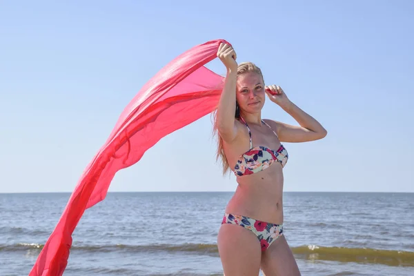 Chica rubia en bikini de pie en la playa y sosteniendo un pañuelo de seda roja. Hermosa mujer joven en un bikini colorido sobre fondo de mar —  Fotos de Stock
