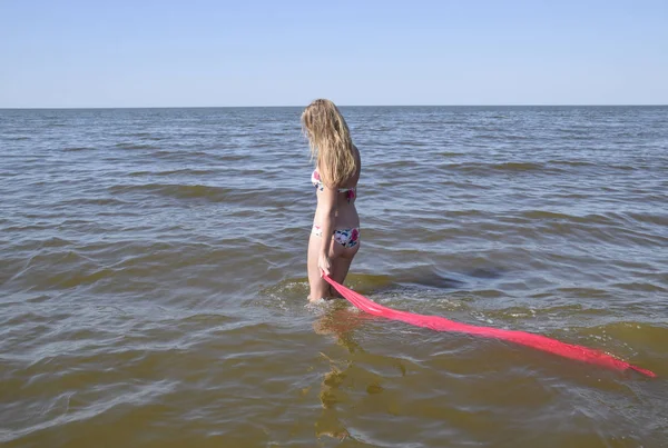 Blond flicka i en bikini står på stranden och håller en röd siden näsduk. Vacker ung kvinna i en färgglad bikini på havet bakgrund — Stockfoto