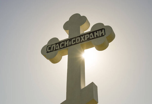 Orthodox cross against the blue sky and the sun. Cross in the backlight of the sun. Symbol of the Christian faith. The inscription on the cross: salvation and preservation