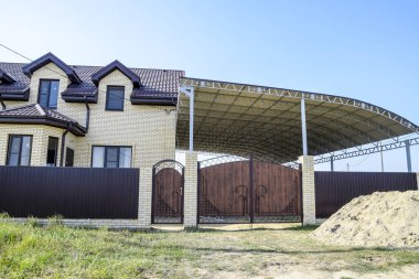 Bir çit ve gates ile tuğla ev. Çelik çerçeve ile büyük bir tente. Görünümü yeni bir İnşaatlı çit ve tuğla ve oluklu metal yapılmış bir ev