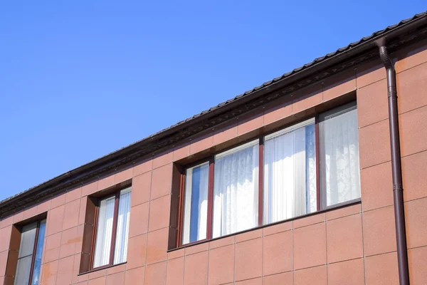 Plastová okna na domě a přepad systému na střeše. Dům s plastovými okny a hnědá střecha z vlnitého plechu — Stock fotografie