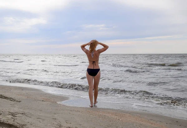 Chica en bikini se encuentra junto al mar, una vista trasera. Un paseo por la playa de la rubia a lo largo de la orilla. La chica va al mar . —  Fotos de Stock