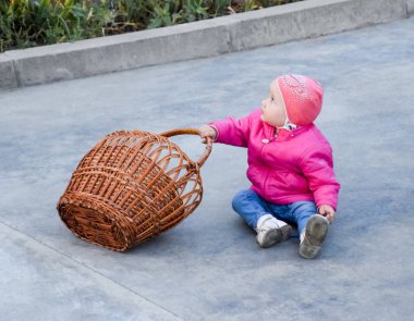 Bir yaşındaki çocuk bir pembe ceket ve şapka beton bir hasır sepet yanında oturur.