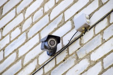 Duvarda ışıklı bir video kamera asılı. Beyaz tuğladan bir duvar ve kablolu bir kamera..