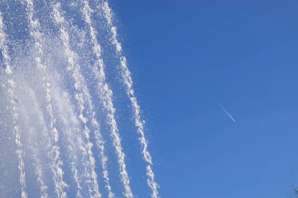 Jets and splashes of water fountain against the blue sky. Condensation track of a jet plane against the sky and a fountain.