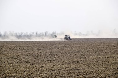 Traktör toprak sahada tırmıklar ve arkasında toz bulutu oluşturur
