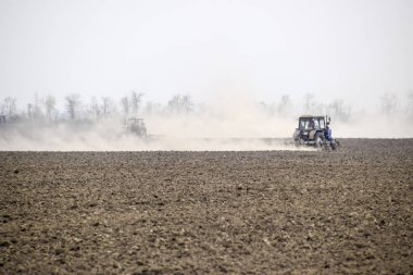 Traktör toprak sahada tırmıklar ve arkasında toz bulutu oluşturur