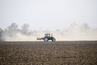 Traktör toprak sahada tırmıklar ve arkasında toz bulutu oluşturur