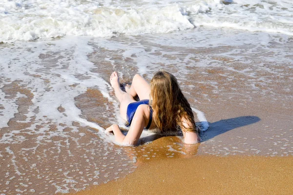 Flicka i en blå baddräkt ligger på havsstranden och slappnar av. Vågorna smeker flickor kroppen — Stockfoto