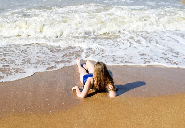 Dívka v modrých plavkách leží na pobřeží a uvolňuje. Vlny pohladí tělo dívky — Stock fotografie