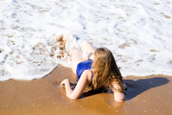 Dívka v modrých plavkách leží na pobřeží a uvolňuje. Vlny pohladí tělo dívky — Stock fotografie