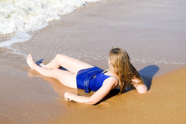 Chica en un traje de baño azul se encuentra en la orilla del mar y se relaja. Olas acarician el cuerpo de las niñas —  Fotos de Stock